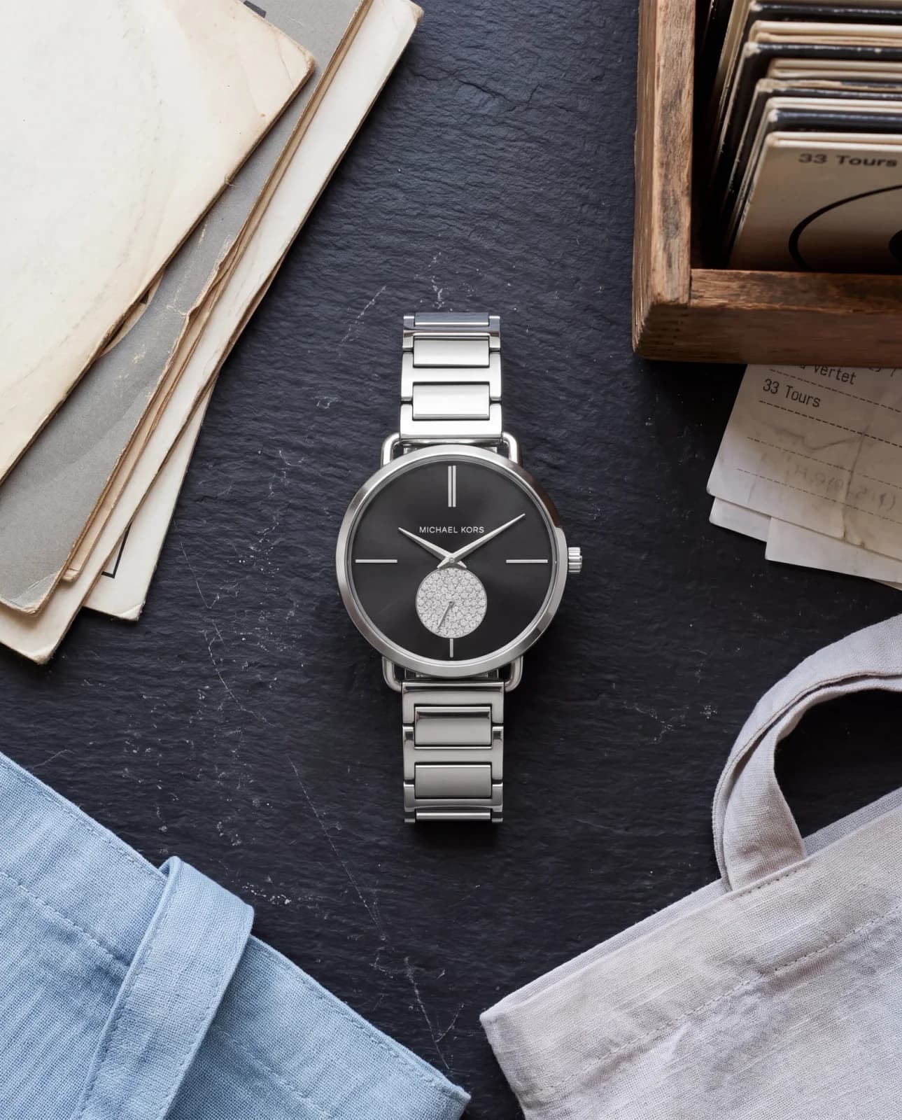 Montre Michael Kors Portia MK3638 à cadran noir et bracelet acier, posée vue du dessus sur un comptoir en ardoise d'un disquaire parisien
Vérification : enviro